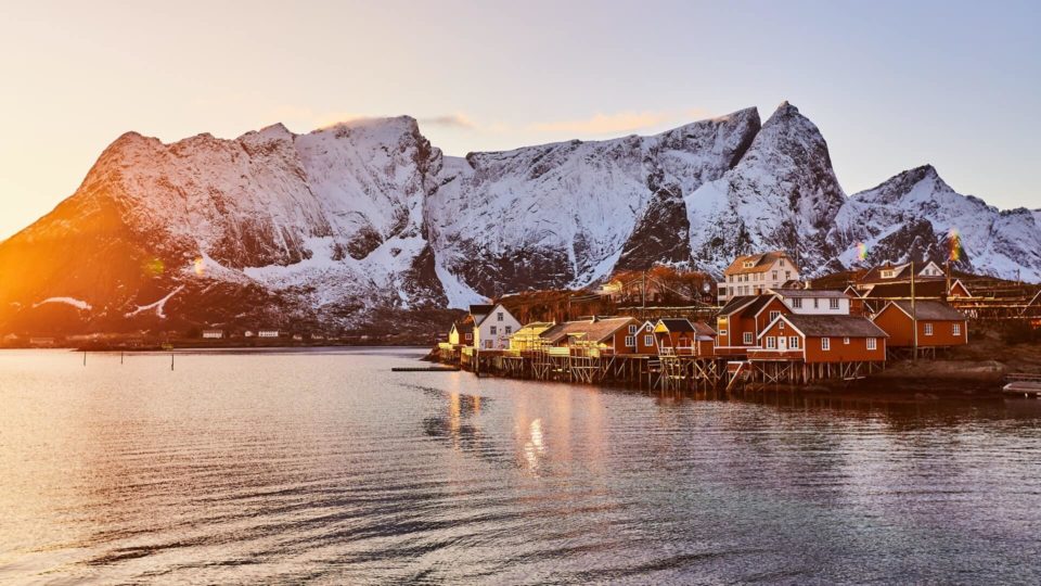 Røde træhuse på pæle ligger langs den rolige havnefront med snedækkede bjerge, der rejser sig i baggrunden under den nedgående sol.