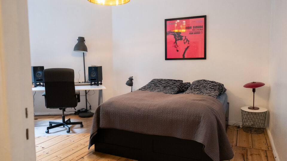 Et ryddeligt soveværelse med en dobbeltseng, skrivebord og stol, computer med højttalere, en gulvlampe, et lille sidebord med en rød lampe og en indrammet plakat på væggen.