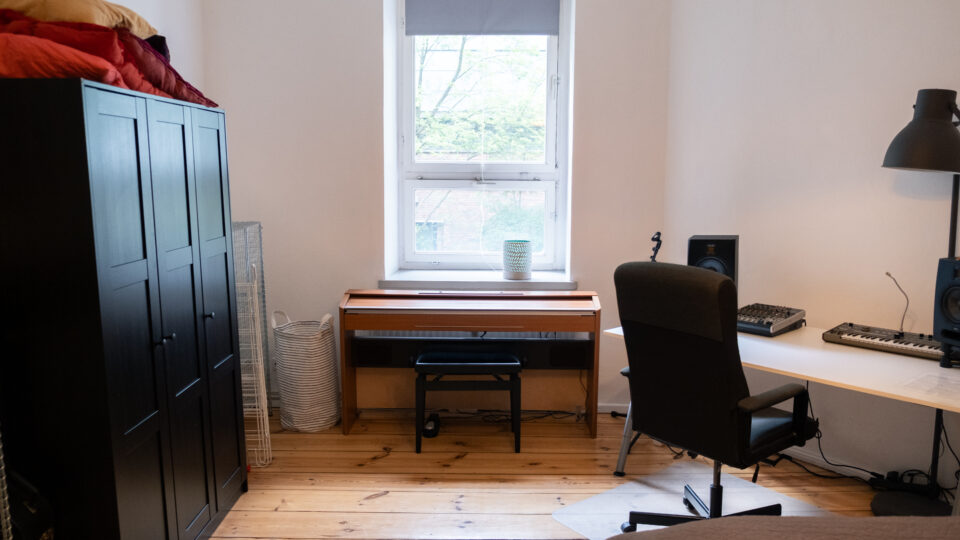 Et ryddeligt soveværelse med et klædeskab, et skrivebord med lydudstyr, et klaver under et vindue og en seng, der er delvist synlig i forgrunden.