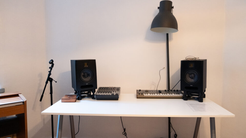Et hvidt skrivebord med to studiemonitorer, en mixer og en keyboard-synthesizer står op ad en hvid væg under en sort gulvlampe.