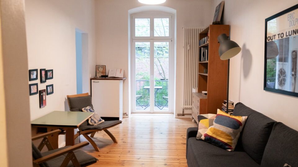 En lille, pæn stue med trægulv, en sofa med puder, to stole ved et grønt bord, bogreoler, en gulvlampe og en vinduesdør, der fører ud til en balkon.