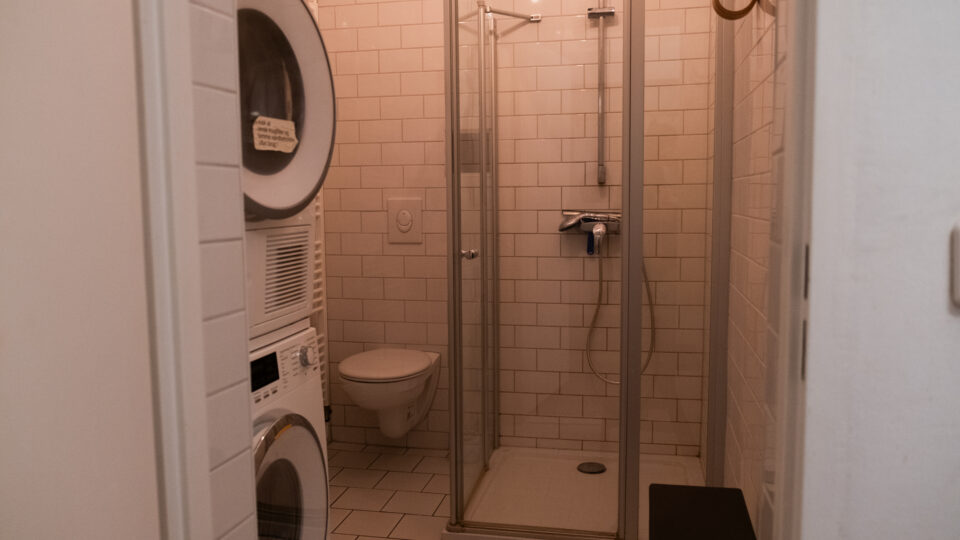 Et badeværelse med glasbruser, væghængt toilet og en stablet vaskemaskine og tørretumbler mod flisebelagte vægge og gulv.