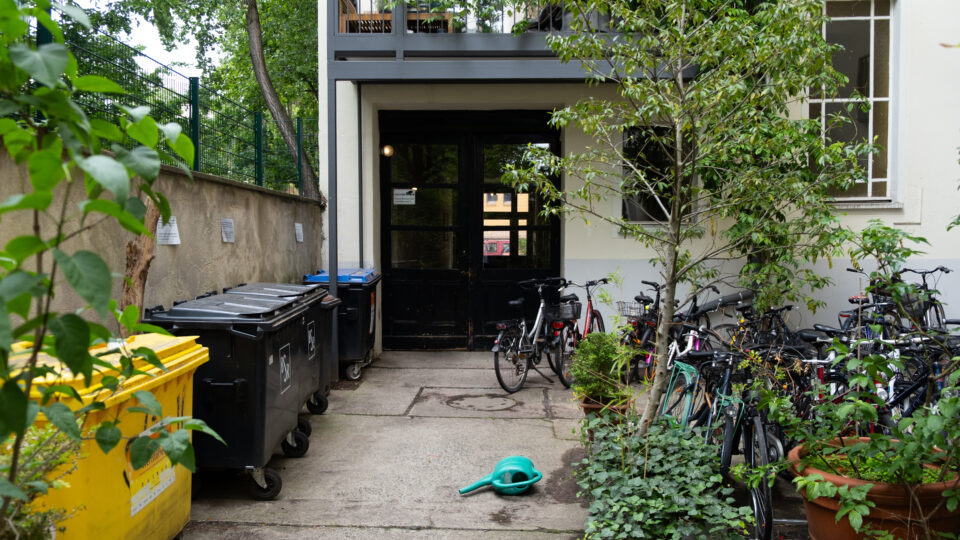 En gårdsplads med cykler parkeret langs højre side, skraldespande til venstre, potteplanter og en grøn vandkande liggende på jorden nær en dobbeltdør til indgangen.