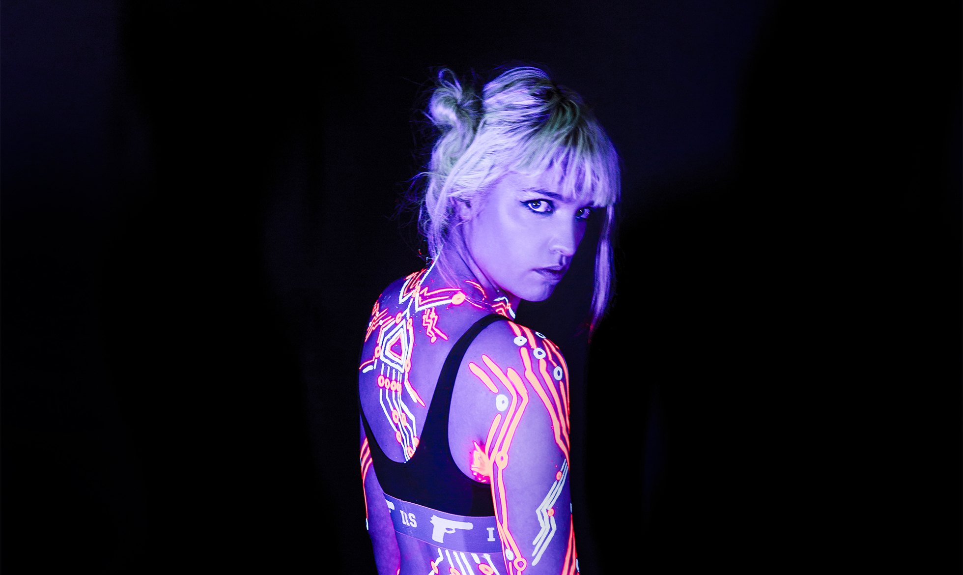 Lydmor, en kvinde i neon-bodysuit, poserer foran en mørk baggrund.