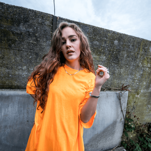 En pige ved navn Amalie Jensen i en orange t-shirt lænet op ad en væg.