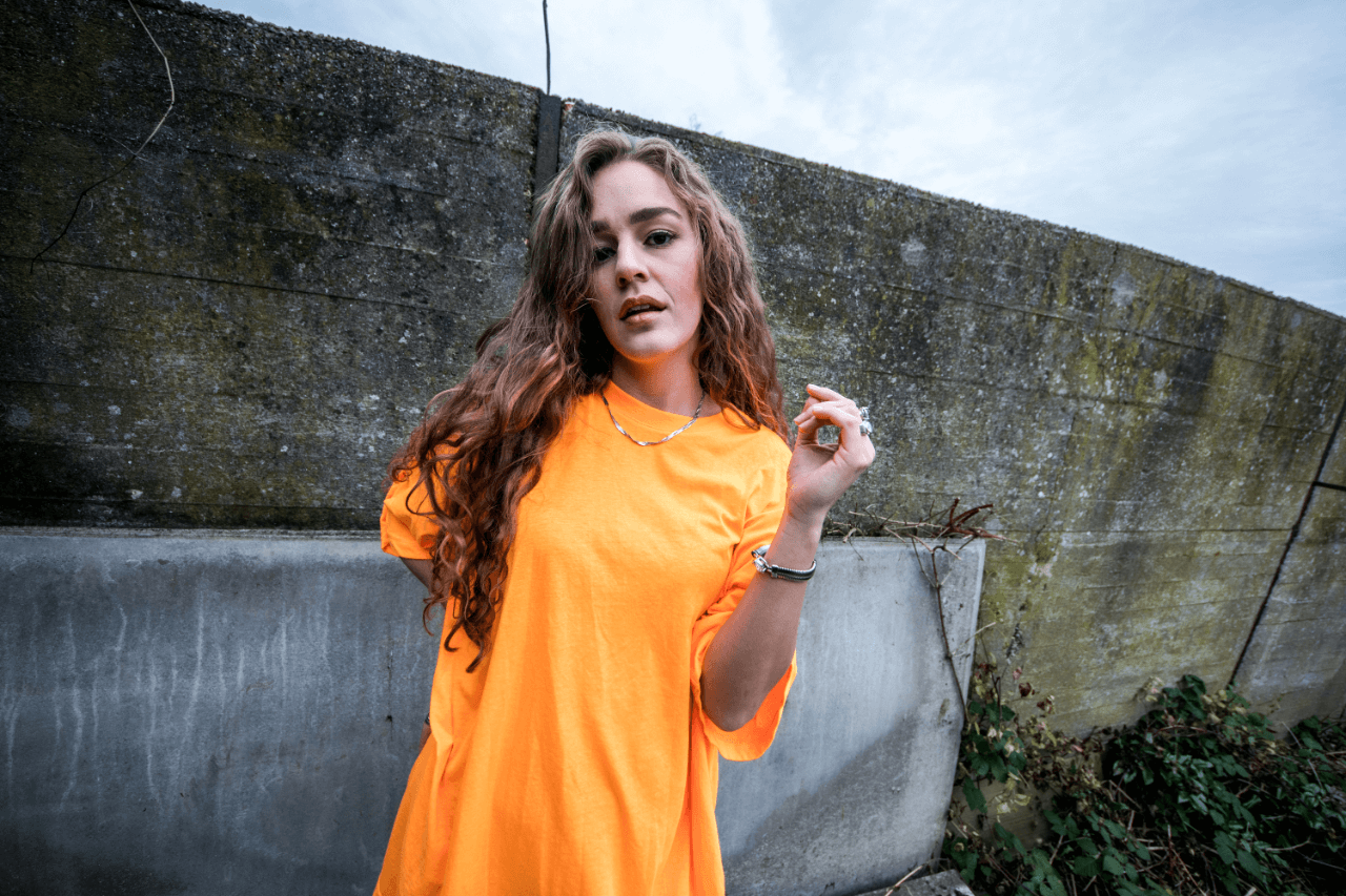 En pige ved navn Amalie Jensen i en orange t-shirt lænet op ad en væg.