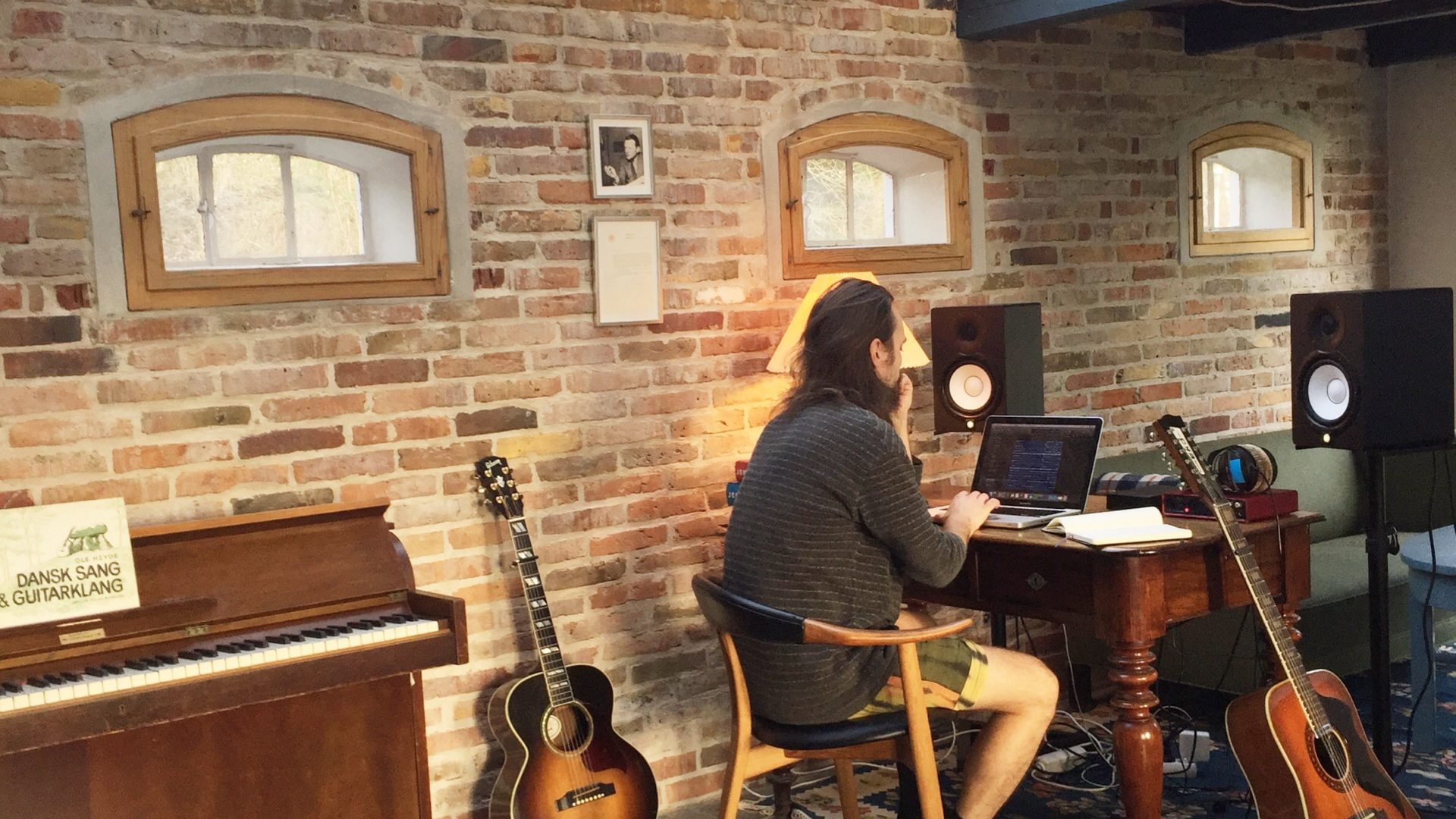 En mand sidder ved et skrivebord i et Lille værelse med en guitar.