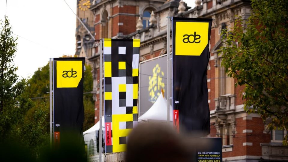 En gruppe mennesker står foran en bygning i Amsterdam med gule og sorte bannere og viser Electronic Fusion-vibes.