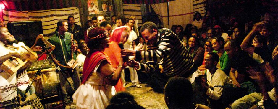 En gruppe mennesker med solbrune ansigt danser i et fyldt lokale.
