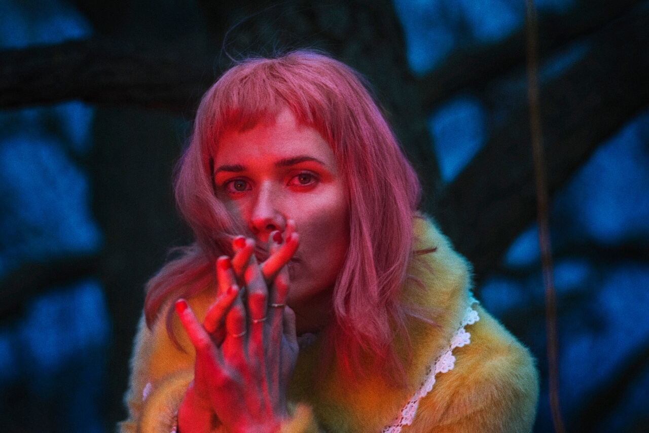 En kvinde med lyserødt hår, kendt som Oh Land, ryger en cigaret i en skov.