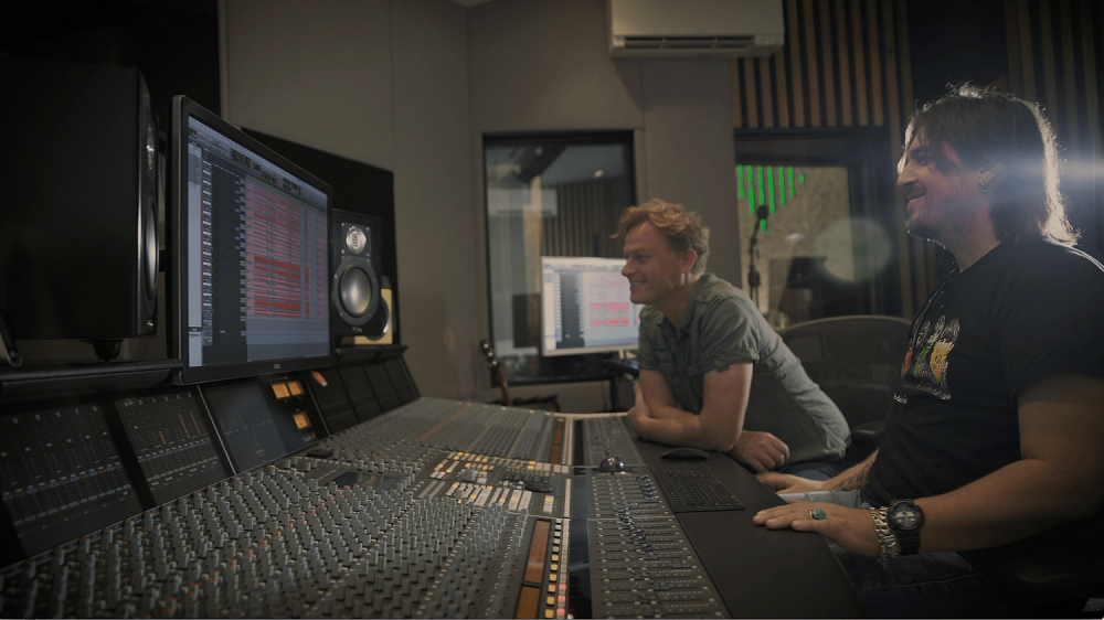 To mænd, der sidder foran et mixebord, lærer online kursus om musikproduktion og perfektionerer deres vokalfærdigheder.