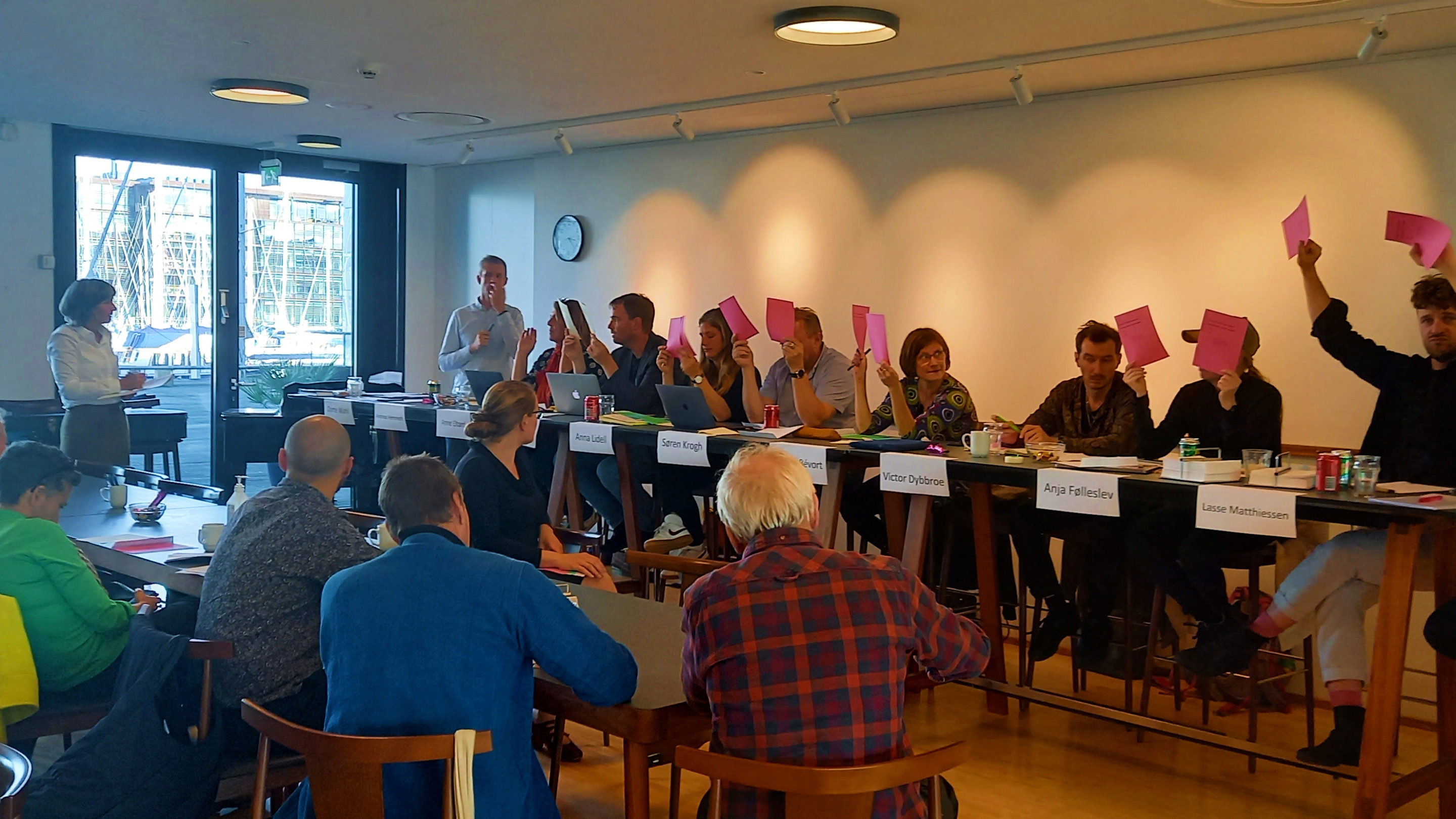 En nyvalgt bestyrelse af folk, der sidder ved et bord og holder lyserødt papir op og viser resultaterne fra generalforsamlingen.