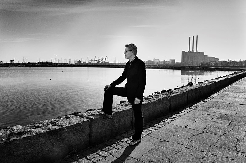 Et sort/hvidt foto med Kjeld Lauritsen lænet op ad en væg nær en vandmasse med en antydning af mystik.