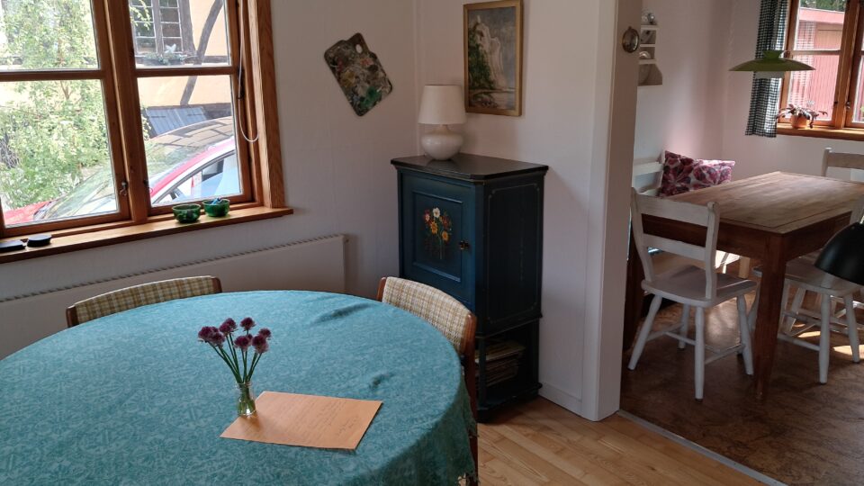 Et rundt bord med en blågrøn dug, en vase med blomster og en mappe står i et solbeskinnet rum. I nærheden står et skab, vægkunst og et spisebord med stole i et tilstødende rum.