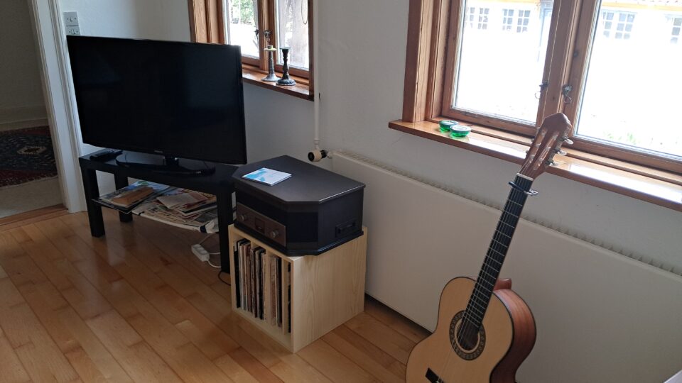 Et stuehjørne med et tv på et stativ, en pladespiller på en træhylde med vinylplader under, og en akustisk guitar på gulvet ved et vindue.