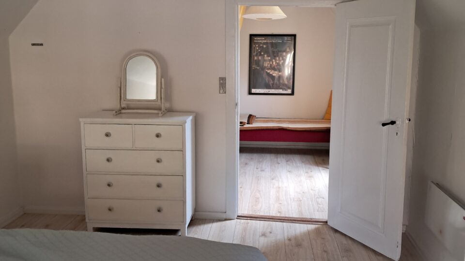 Et enkelt soveværelse med en seng, en hvid kommode og et spejl med en åben dør, der fører ind til et andet rum med en rød daybed og indrammet vægkunst.