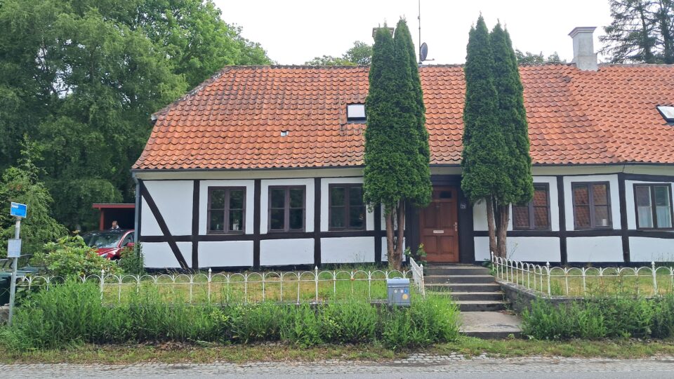 Et bindingsværkshus med rødt tegltag, to høje træer ved trappen og et hvidt hegn ud mod vejen.