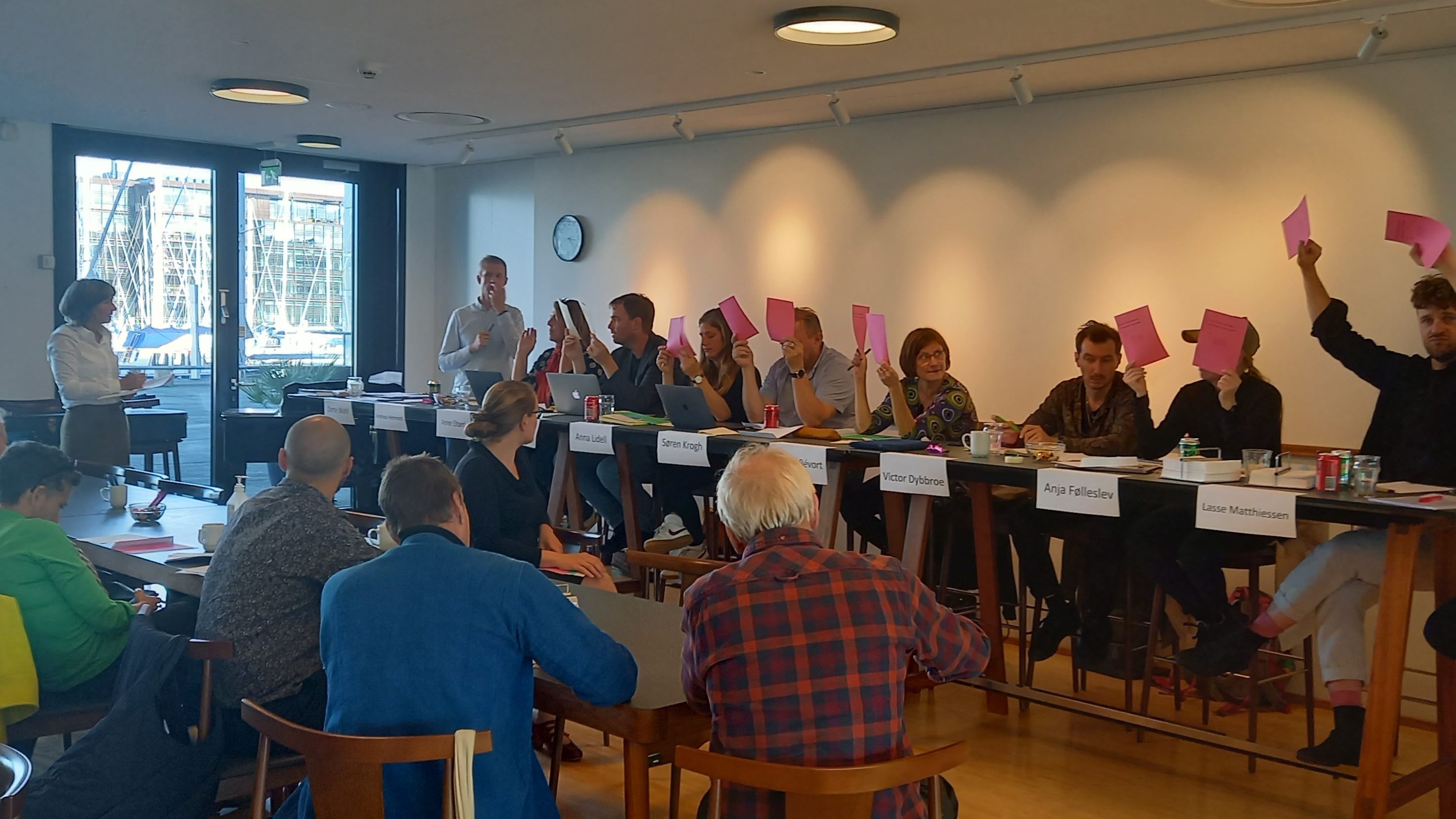 En gruppe generalforsamlingsdeltagere sidder ved et bord og holder lyserødt papir op.