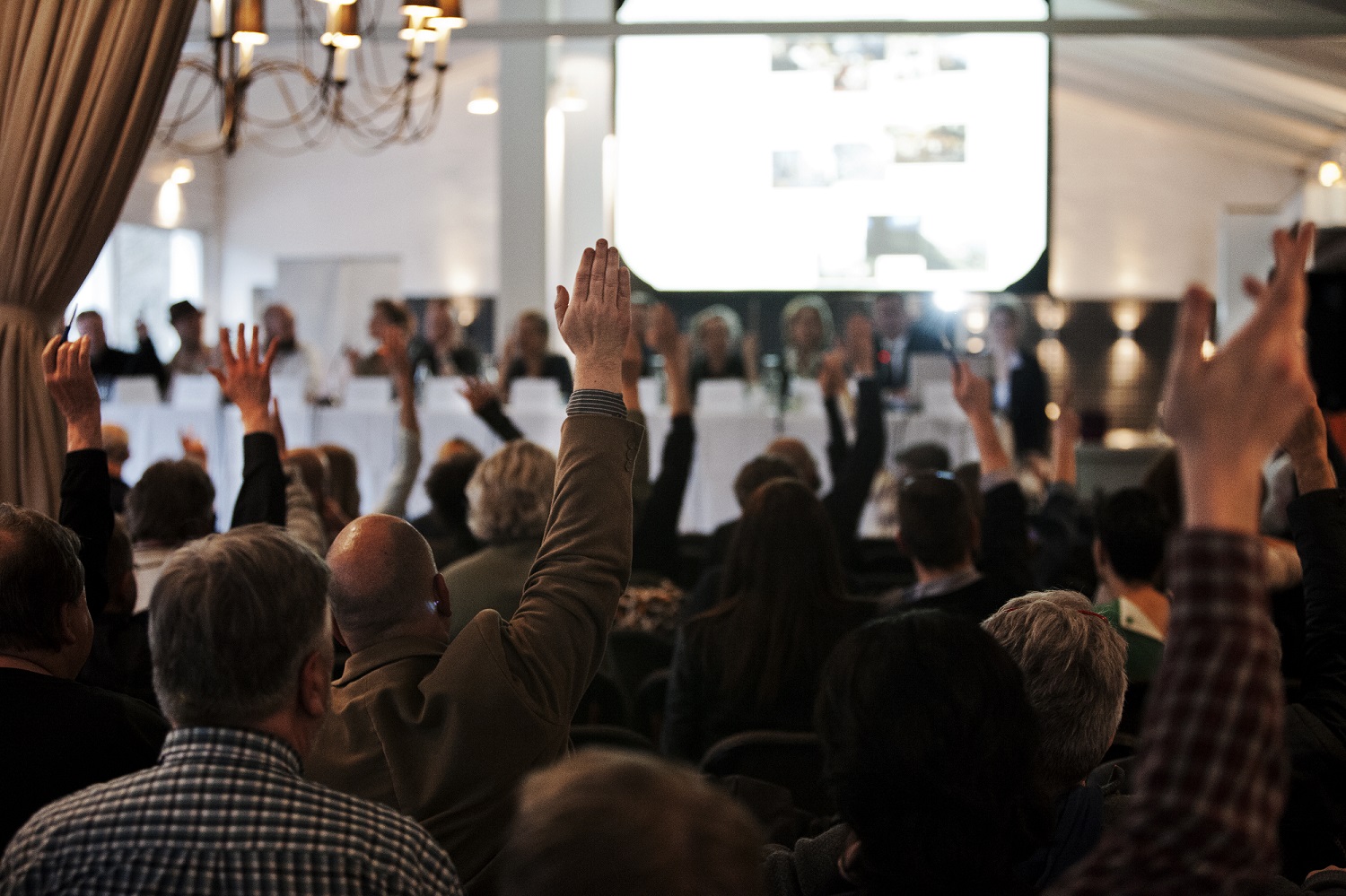 En gruppe mennesker, der deltager i en ekstraordinær generalforsamling, rækker hænderne op i et lokale.