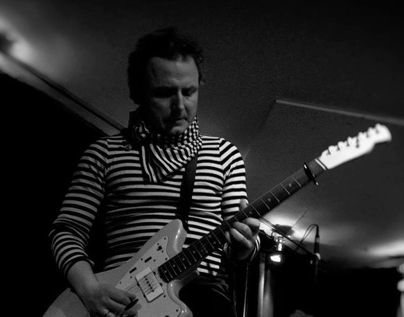 En mand iført en stribet skjorte og et tørklæde spiller på en elektrisk guitar på scenen, i svagt lys og med en mørk baggrund.