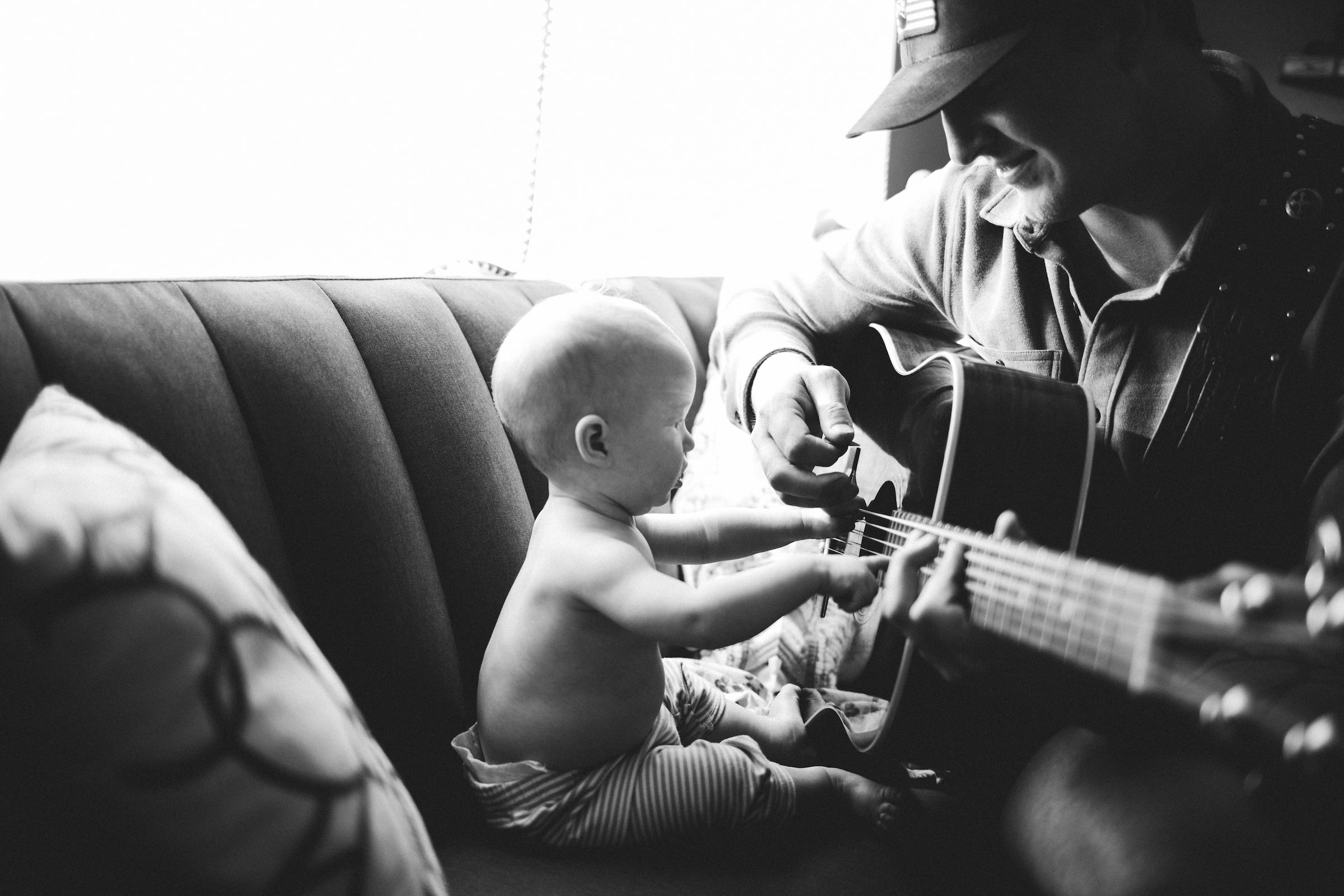 En musikforældre spiller guitar med en baby på en sofa under en online workshop.