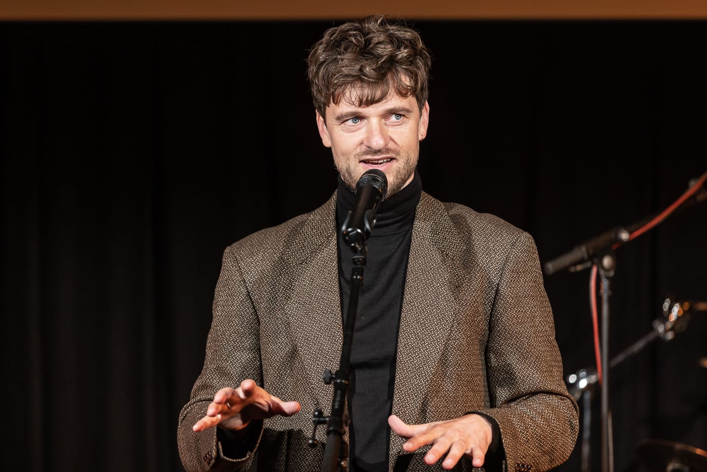 En mand i brun jakke, der står foran en mikrofon og holder en kulturpolitisk tale.