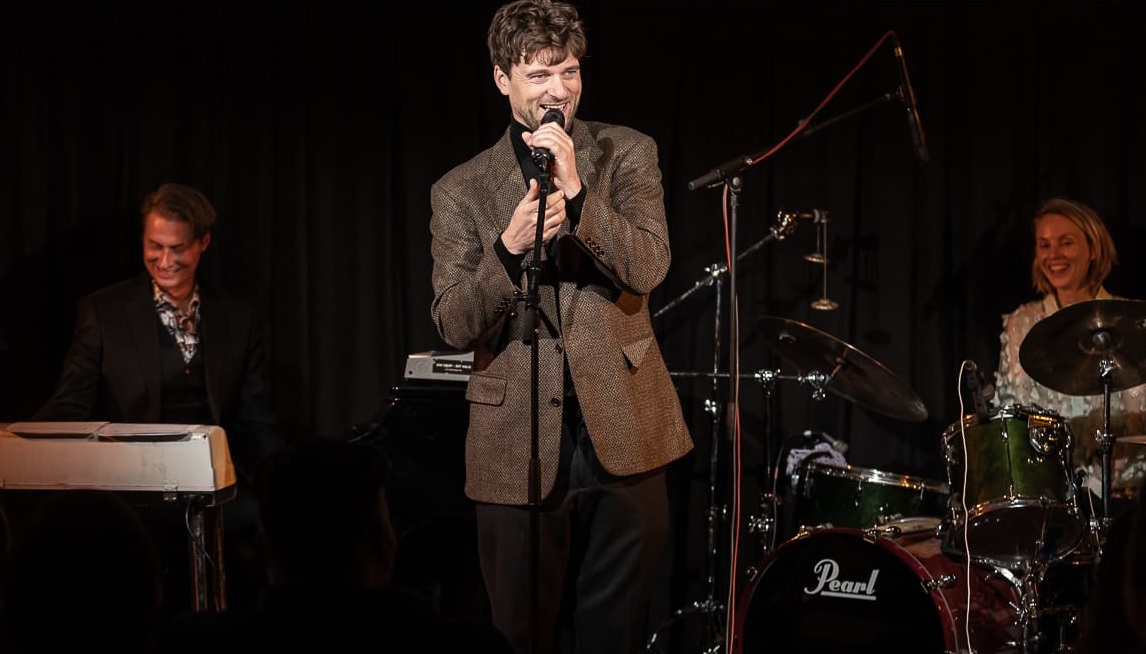 En mand synger i en mikrofon foran en gruppe musikere og skaber en os alle-forestilling.