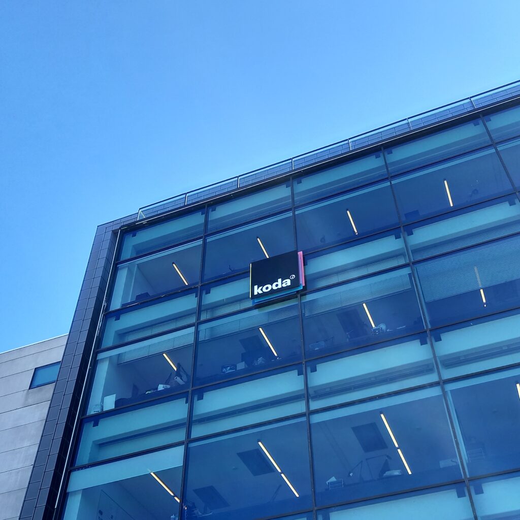 Deltag i Kodas generalforsamling digitalt foran en glasbygning med en blå himmel i baggrunden.