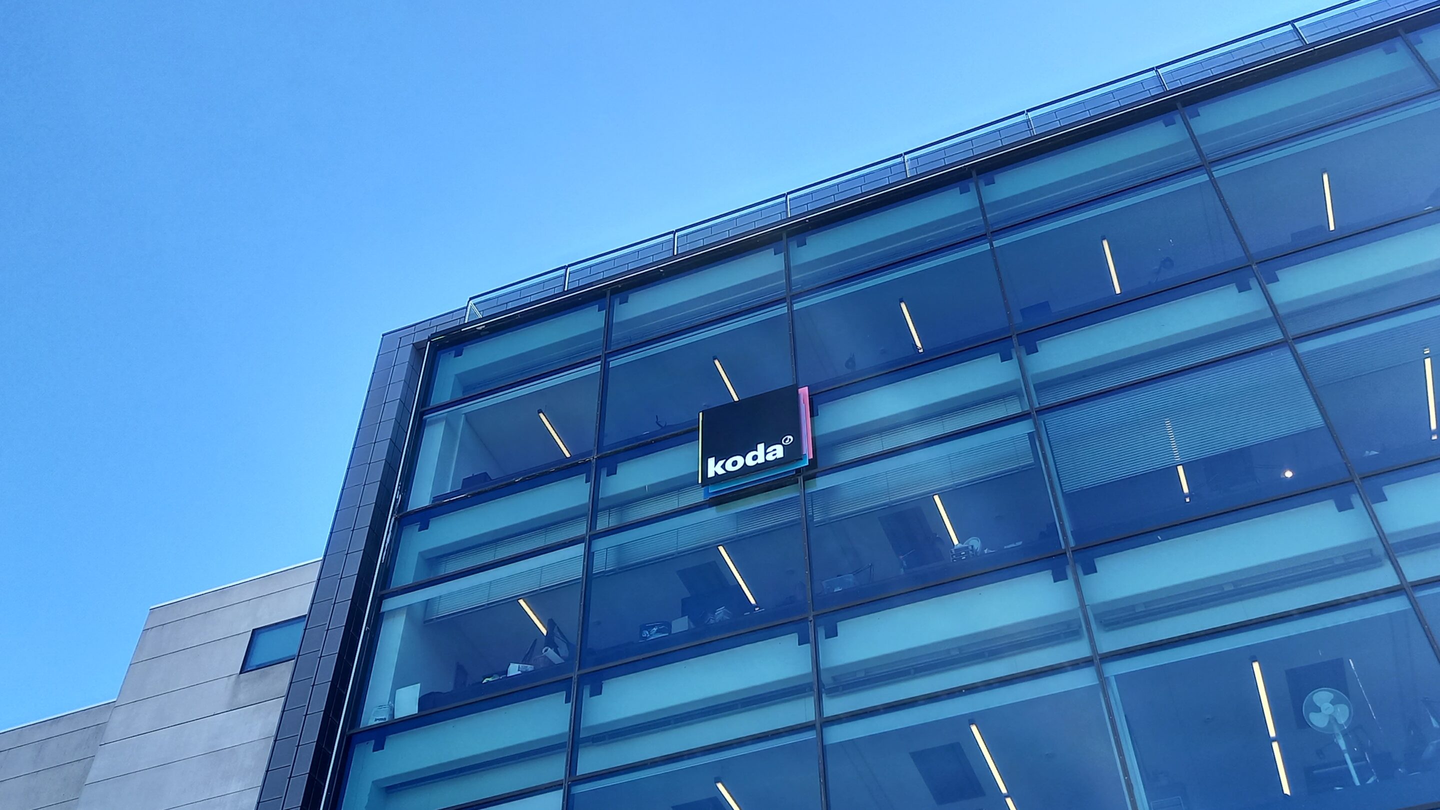 Deltag i Kodas generalforsamling digitalt foran en glasbygning med en blå himmel i baggrunden.
