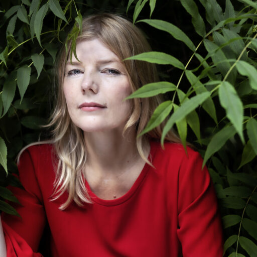 Anna Brønsted, en kvinde i rød top, gemmer sig bag nogle grønne blade.