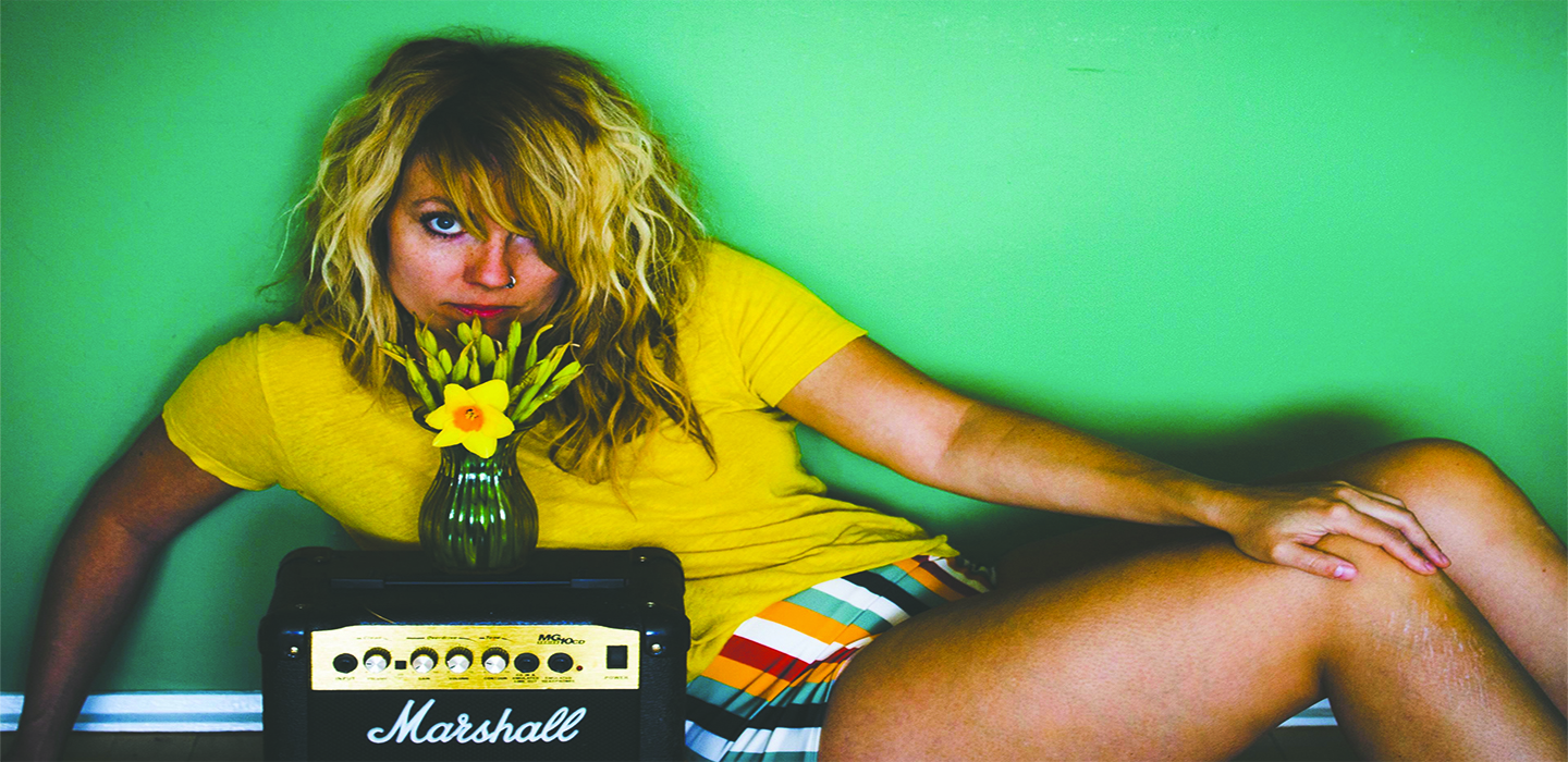 En kvinde med GrownUp Gold-hår, der sidder på toppen af en guitarforstærker.