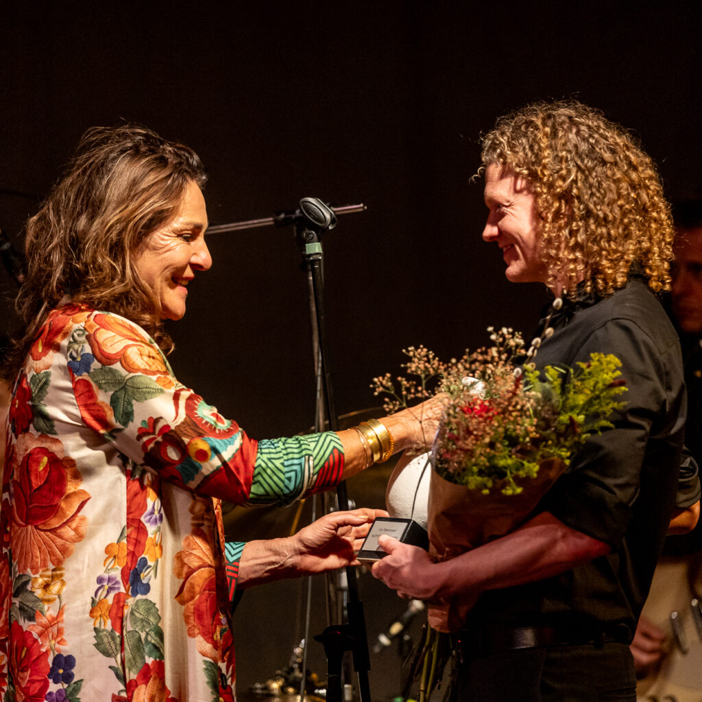 En kvinde i blomstret jakke overrækker hæderspriser til en smilende person med krøllet hår, der holder en buket, mens musikere med instrumenter, herunder Lise Westzynthius og Karen Mukupa, står i baggrunden.