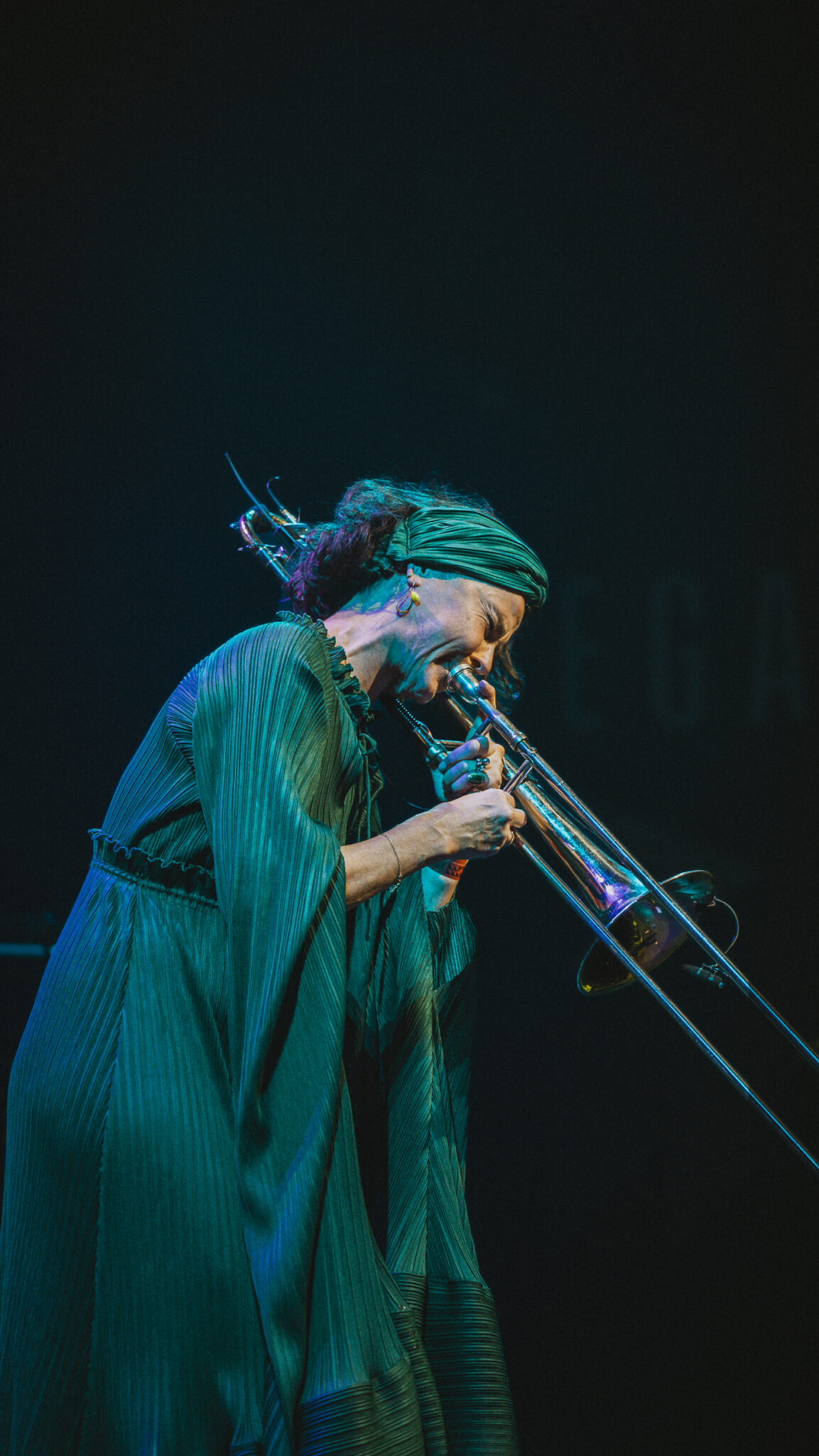 En musiker iført grønt outfit og headwrap spiller trombone på scenen mod en mørk baggrund, lidenskabeligt støttet af et arbejdslegat, der giver næring til deres kreative rejse.