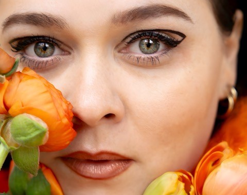 Nærbillede af en person med fed øjenmakeup, omgivet af orange blomster, der viser en sangerkrop, der udstråler selvtillid.
