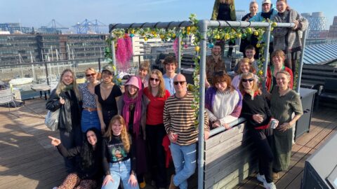 En gruppe mennesker poserer sammen på en tagterrasse dekoreret med en blomsterbue med et bybillede i baggrunden. Nogle står i ånden fra forår 2024, mens andre sidder under den klare himmel og fejrer deres samarbejdskraft midt i en betagende byudsigt.