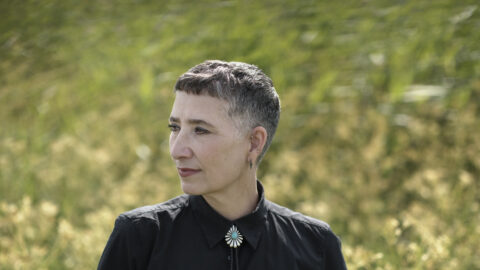 Trinelise Væring, med kort hår og et boloslips med sin sorte skjorte, står på en mark med en luft af Medlem i fokus og kigger eftertænksomt til siden.