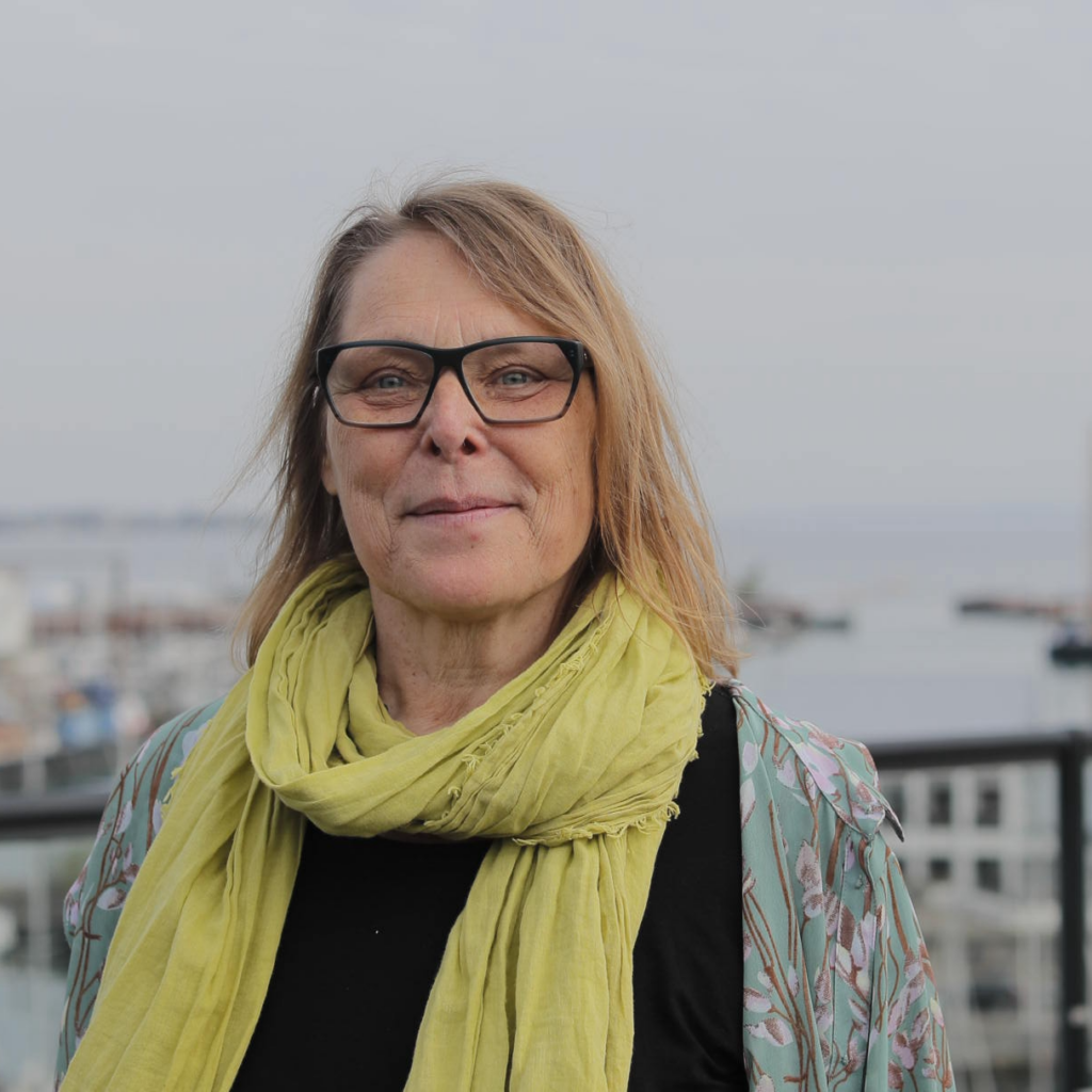 En kvinde med skulderlangt blond hår, briller og et lysegrønt tørklæde står udendørs nær en havnefront med både og bygninger i baggrunden.