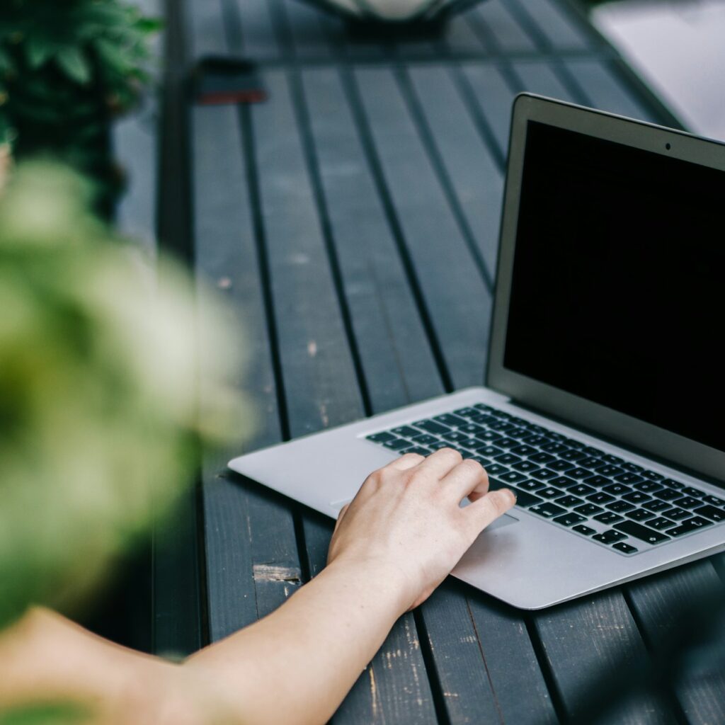 En persons hånd hviler på tastaturet på en åben bærbar computer på et mørkt træbord med grønne blade delvist synlige i forgrunden.