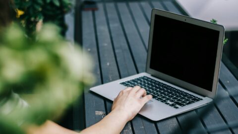 En persons hånd hviler på tastaturet på en åben bærbar computer på et mørkt træbord med grønne blade delvist synlige i forgrunden.