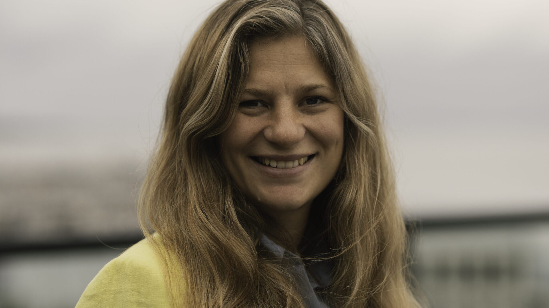 En kvinde i en gul jakke smilende foran en organisationsbygning.