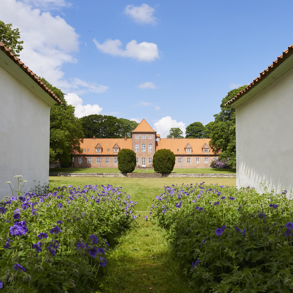 Udsigt mellem to hvide bygninger med en sti omkranset af lilla blomster, der fører til en stor murstensbygning med rødt tag i baggrunden under en blå himmel med skyer.