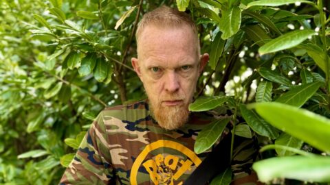 En mand med et alvorligt udtryk, iført camouflageskjorte og med en banjo i hånden, står blandt tæt, grønt løv.