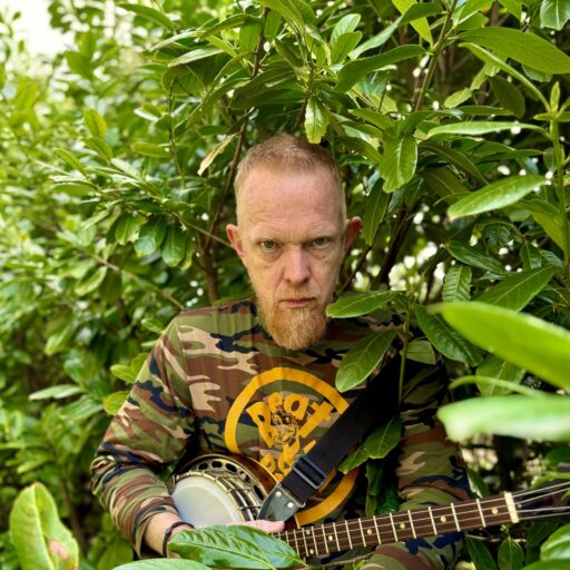 En mand med et alvorligt udtryk, iført camouflageskjorte og med en banjo i hånden, står blandt tæt, grønt løv.