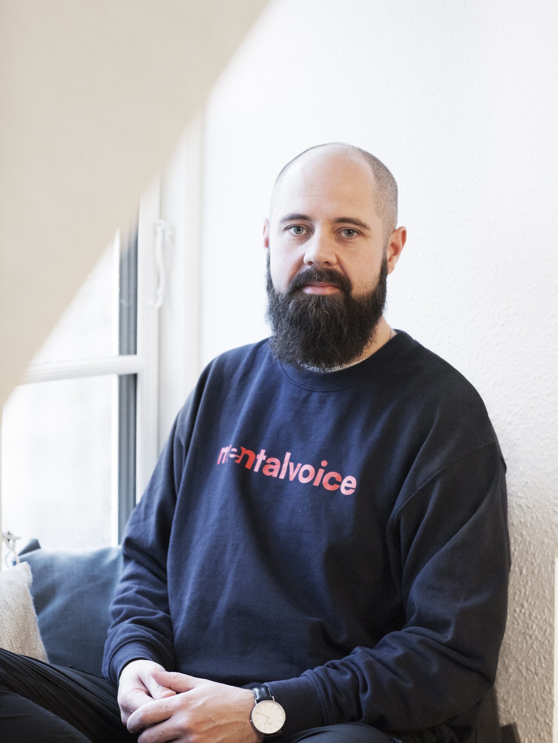 En skægget mand med et barberet hoved sidder indendørs ved et vindue iført en mørk sweatshirt med ordet "mentalvoice" trykt på og et ur på sit venstre håndled.