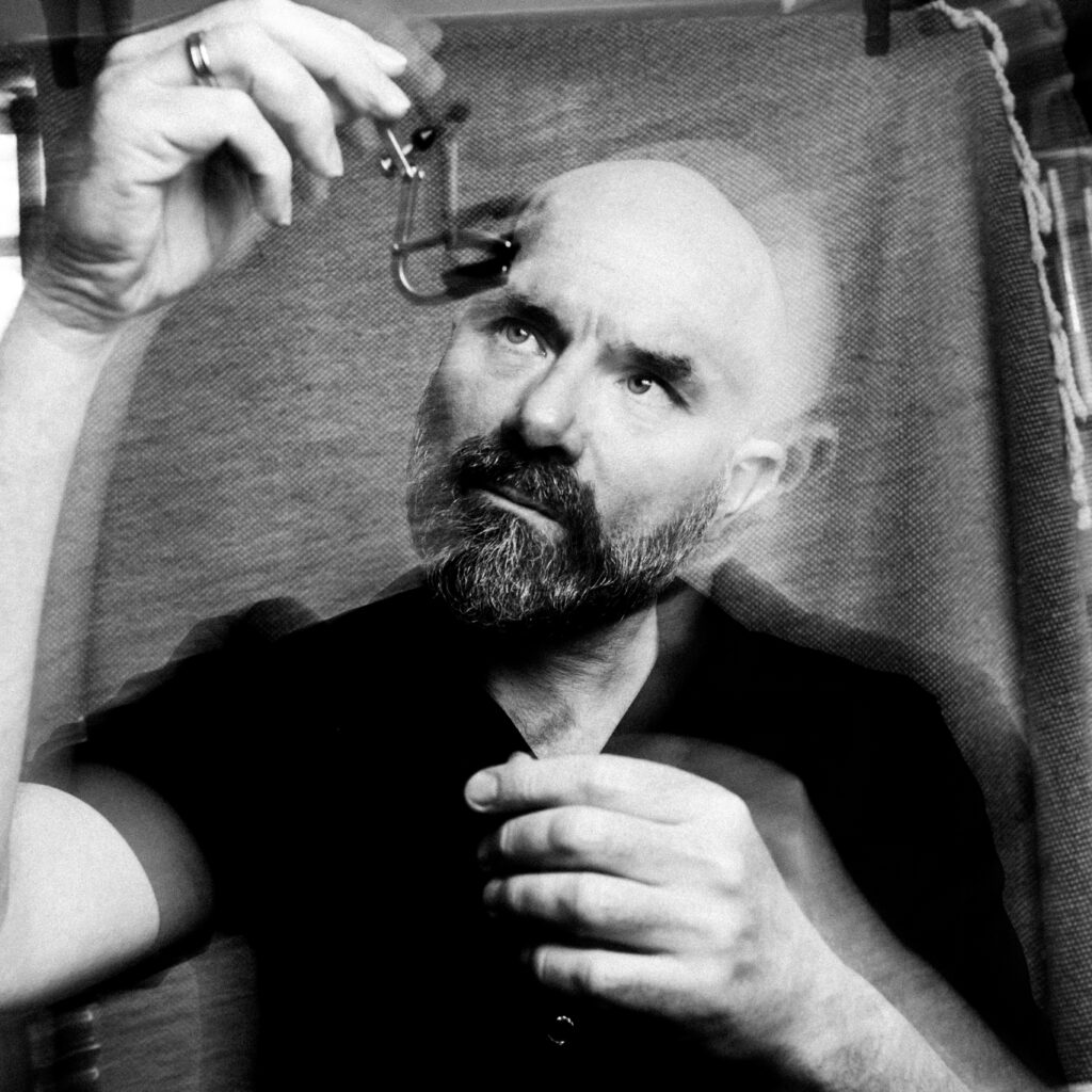 Sort-hvidt foto af en skægget mand, der holder en lille genstand op mod lyset, med en dobbelteksponeringseffekt, der skaber en fornemmelse af bevægelse.