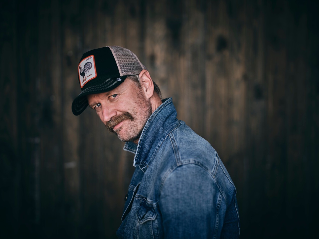 Mand med overskæg iført denimjakke og truckerhat, der står på en mørk træbaggrund og kigger ind i kameraet.