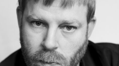 Sort-hvidt portræt af en mand med lyst hår og skæg, der ser direkte ind i kameraet med et neutralt udtryk og er iført en mørk skjorte.