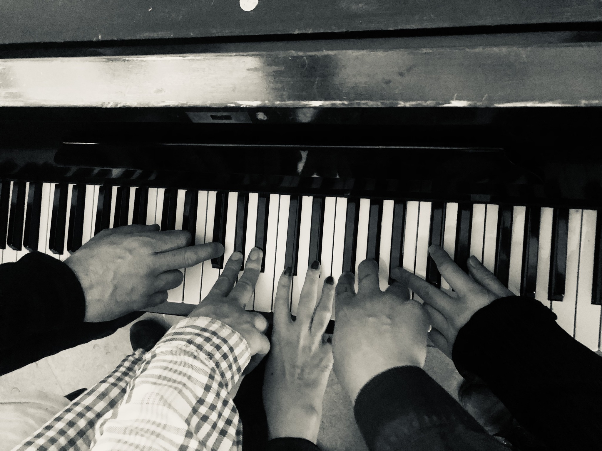 Fire par hænder, der tilhører mennesker i forskellige aldre, spiller sammen på et klaverkeyboard på et sort-hvidt foto.