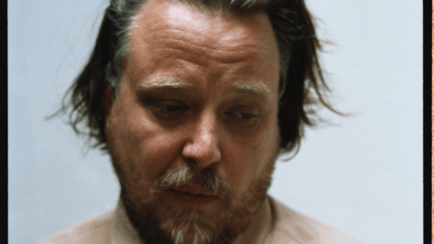 En mand med lys hud, pjusket mørkeblondt hår og skæg kigger ned, mens han er iført en beige skjorte med knapper på en almindelig lys baggrund.