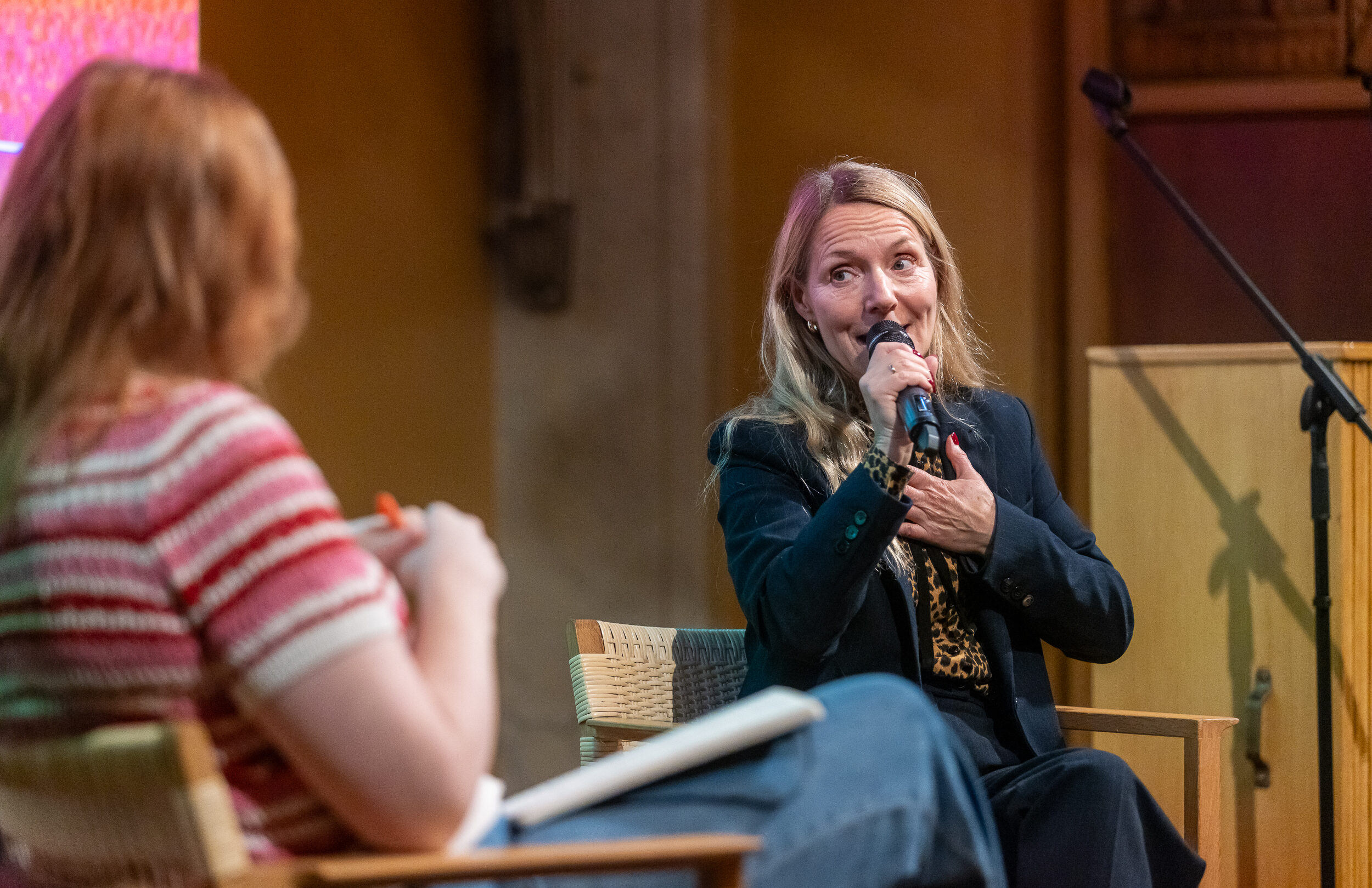 En kvinde taler i en mikrofon, mens hun sidder på scenen med front mod en anden siddende person, der holder en notesbog og en kuglepen.
